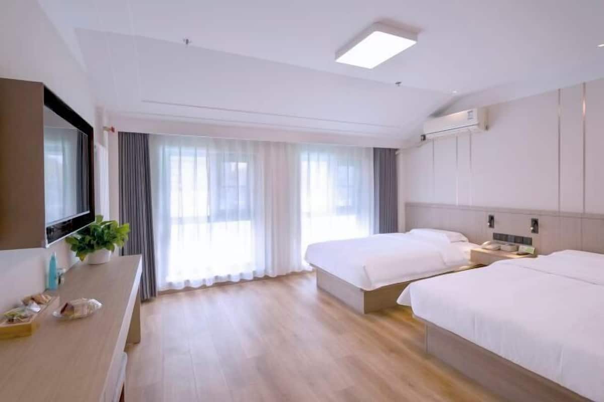 A hotel room with two beds, a desk with a plant, a TV, and a large window with curtains.