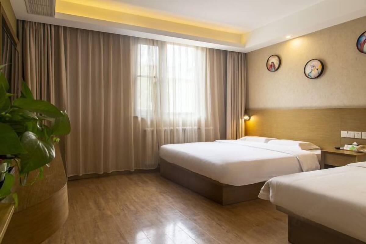 A hotel room with two beds, a large window with curtains, a bedside table with a lamp, and a wall-mounted clock.