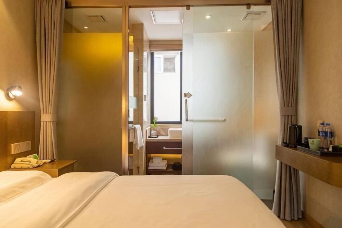 A hotel room with a bed, a nightstand, a mirror, a window with curtains, and a bathroom visible through a door.