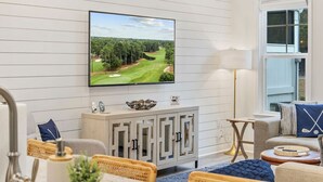 A modern living room with a flat-screen TV mounted on the wall, a wooden cabinet with decorative items, a comfortable seating area with a sofa and chairs, and a window with a view of trees. - The Par-fect Stay at Barefoot Resort (North Myrtle Beach)