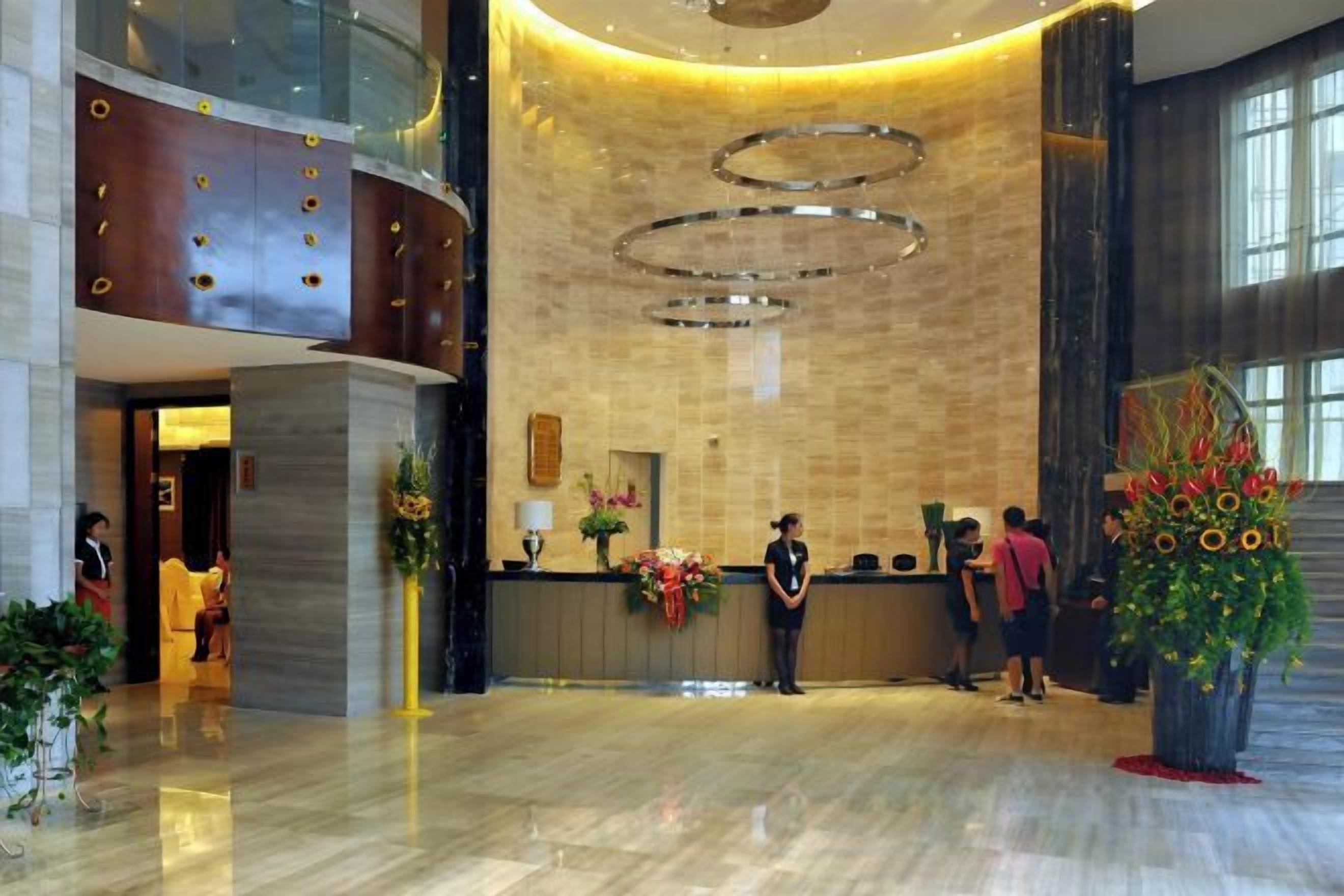 A hotel lobby with a reception desk, a large plant, and a modern chandelier.