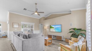 A spacious living room with a grey sofa, wooden chairs, a ceiling fan, and a large TV on the wall. - Coastal Nook: heated outdoor pool, up to 8 guests, 3 bedrooms, & game room (Cape Coral)