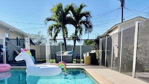 A swimming pool with inflatable unicorn and rainbow floats, surrounded by a fenced area with palm trees and a screened patio. - Coastal Nook: heated outdoor pool, up to 8 guests, 3 bedrooms, & game room (Cape Coral)