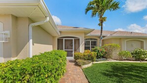 A residential house with a paved driveway, a front door, and a small garden with various plants and a palm tree. - Coastal Nook: heated outdoor pool, up to 8 guests, 3 bedrooms, & game room (Cape Coral)