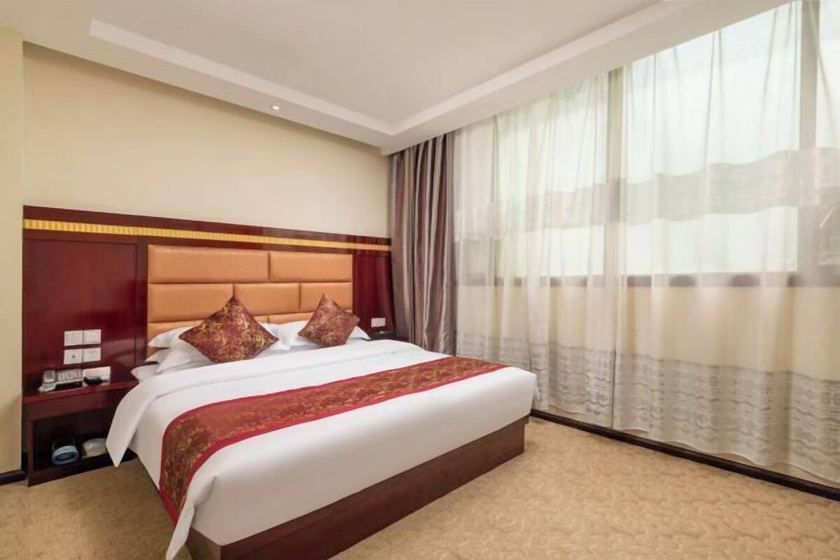 Hotellihuone, jossa on suuri sänky, puinen pääty, yöpöytä ja ikkuna, jossa on läpikuultavat verhot.