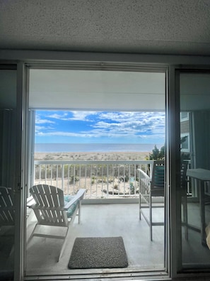 A balcony with a view of the beach, a chair, a table, and a doormat.