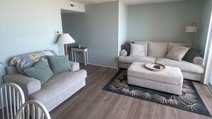 A modern living room with a grey sofa, a smaller grey sofa, a coffee table, a dining table with chairs, and a patterned rug. - NEW Beautiful Ocean Front Condo with Private Balcony and Beach Access (Ocean City)