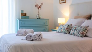 Un lit bien fait, agrémenté de serviettes pliées, d’un coussin à motifs floraux, d’un vase contenant des plantes séchées et d’une commode bleue, dans une chambre d’hôtel.