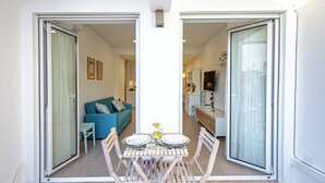 Un coin repas avec une table dressée pour deux personnes, un canapé en arrière-plan et une cuisine visible à travers une porte ouverte.