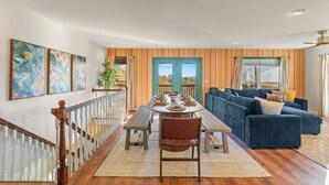 Un vaste espace de vie doté d’un plancher en bois, d’une table à manger pour six personnes, d’un canapé d’angle bleu et d’un escalier avec des rampes blanches.