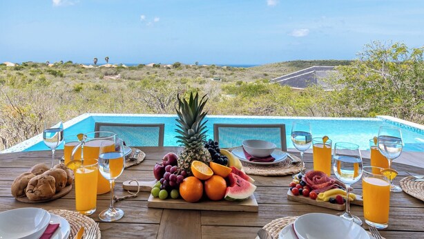 A dining table with a fruit platter, glasses of orange juice, and a view of a swimming pool and landscape. - Villa Kas Di Lotte, 3-bedroom villa in marvelous Coral Estate with AC, WiFi (Sint Willibrordus)