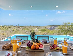 A dining table with a fruit platter, glasses of orange juice, and a view of a swimming pool and landscape. - Villa Kas Di Lotte, 3-bedroom villa in marvelous Coral Estate with AC, WiFi (Sint Willibrordus)