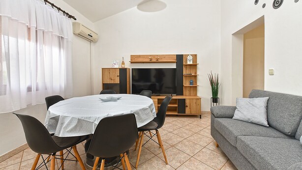 A living room with a grey sofa, a round dining table with four chairs, a TV unit with shelves, and a potted plant. - Apartment 1858/143214 (Istria - Valbandon), 1000m from the beach (Valbandon)