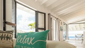A modern living room with a beige sofa, a teal pillow with "relax you're on beach time" text, large windows, and a view of the outdoors. - Villa Neshi, Spacious 5-bedroom villa in charming Cas Abou Resort with AC, WiFi (Willemstad)