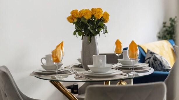 A dining table set with white plates, cups, and glasses, a vase with yellow flowers, and a modern chair design. - Converted Victorian Bakery (London)