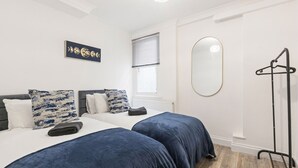 A bedroom with two beds, a radiator, a window with blinds, a wall-mounted mirror, and a clothing rack. - Converted Victorian Bakery (London)