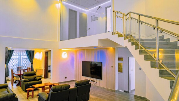 A spacious living area with a flat-screen TV, comfortable seating, a staircase, and a view of the upper floor. - Villa - Sleeps 8 - Parking - Balcony (Dar es Salaam)