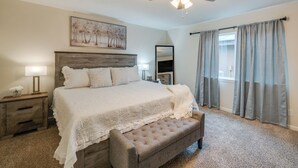 A bedroom with a large bed, a wooden headboard, a ceiling fan, a bedside table with a lamp, a bench, and a window with curtains. - 10 Mi to Six Flags: Pet- & Family-Friendly Retreat (Grand Prairie)