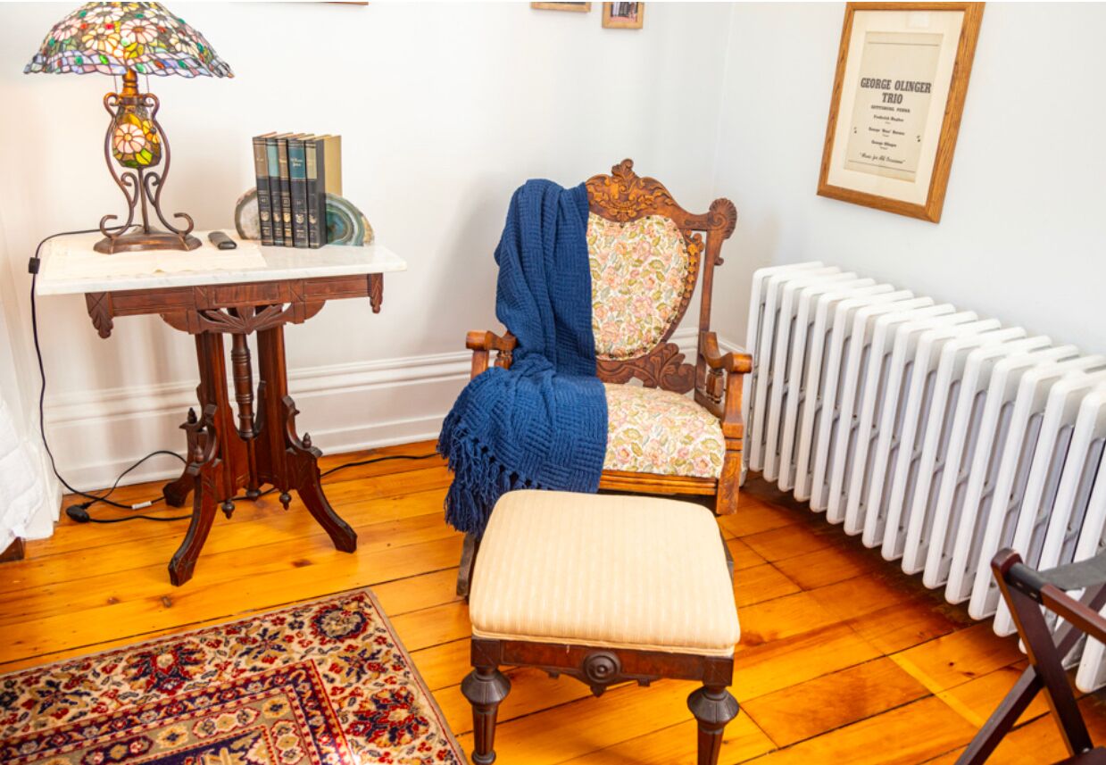 A room with a wooden chair, a small table with a lamp, a radiator, and a framed certificate on the wall.