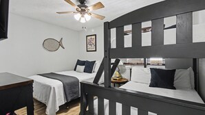 Habitación con literas, ventilador de techo, escritorio y cama con manta blanca y gris.
