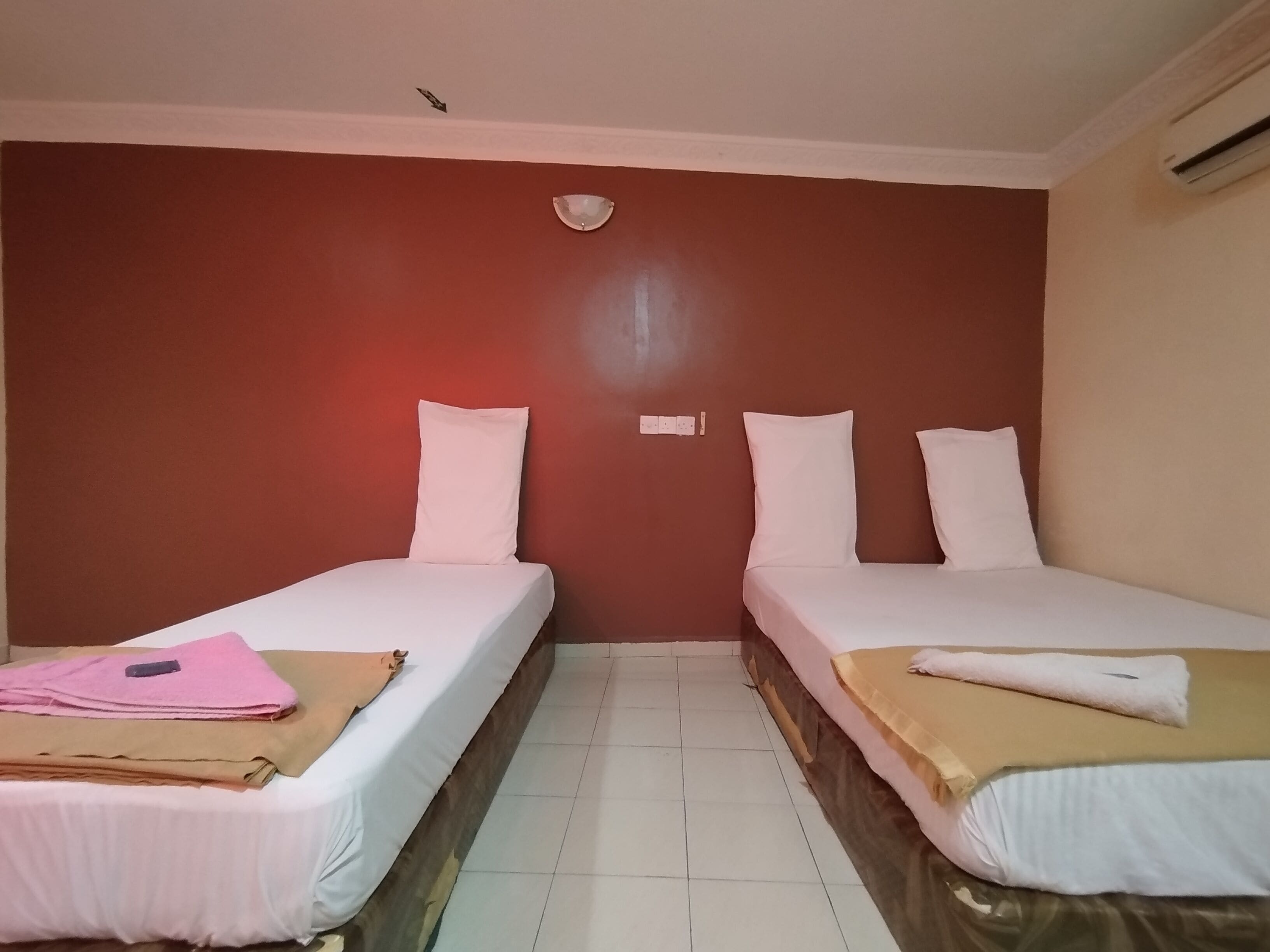 Une chambre d’hôtel avec deux lits simples, chacun équipé de draps et d’oreillers blancs, une petite table avec une serviette rose et un tissu marron, et un climatiseur fixé au mur.