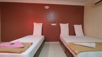 Une chambre d’hôtel avec deux lits simples, chacun équipé de draps et d’oreillers blancs, une petite table avec une serviette rose et un tissu marron, et un climatiseur fixé au mur.