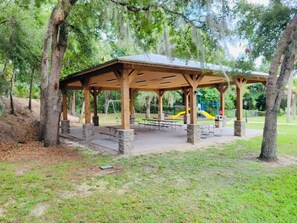 Ein überdachter Pavillon mit Picknicktischen und Bänken, umgeben von Bäumen und einem Spielplatz mit einer Rutsche.