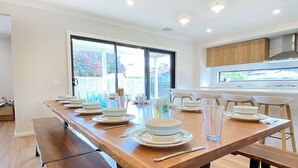 Een moderne eetruimte met een lange houten tafel, gedekt voor een maaltijd, met banken aan één kant en een keuken op de achtergrond.