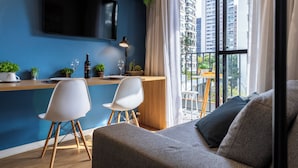 A modern living room with a sofa, two white chairs, a wooden dining table, a TV mounted on the wall, and a view of tall buildings through large windows. - Piscina-AC-Academia-Brazilian Corner (São Paulo)