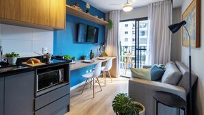 A modern kitchen with a built-in oven, a dining area with a table and chairs, a sofa, and a large window with a view of buildings. - Piscina-AC-Academia-Brazilian Corner (São Paulo)