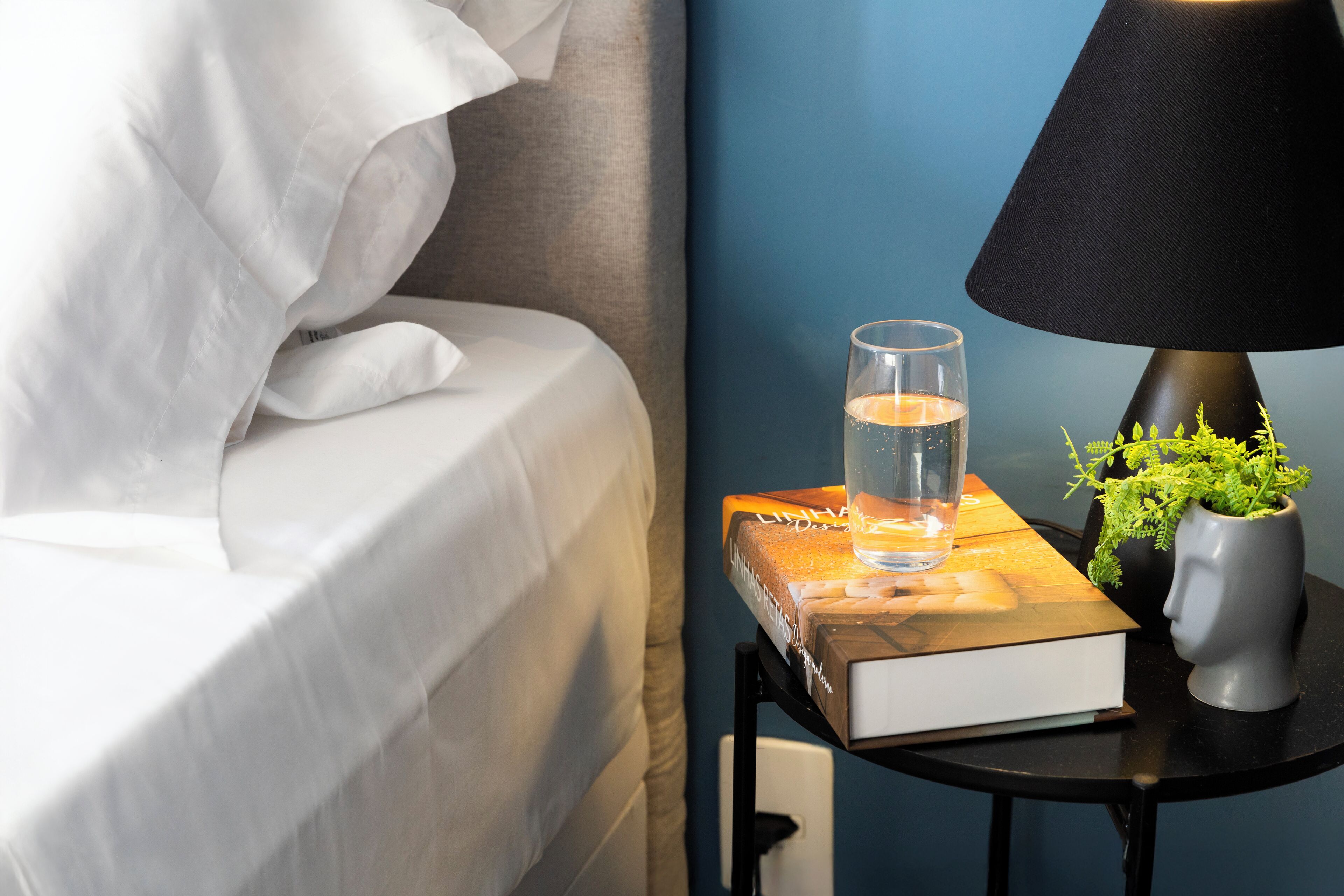 A neatly made bed with white linens, a bedside table with a book, a glass of water, a black lamp, and a small potted plant.