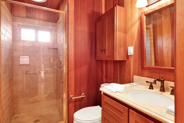 Un baño con cabina de ducha de vidrio, muebles de madera, inodoro blanco y lavabo con grifo antiguo.