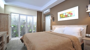 A bedroom with a large bed, a wooden wardrobe, a bedside table, a nightstand, a painting on the wall, and a sliding glass door leading to a balcony with potted plants. - Dasinem Family Homestay (Pogung Lor)