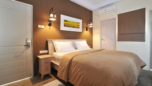 A bedroom with a large bed, two bedside lamps, a nightstand, a painting, and a window with a blind. - Dasinem Family Homestay (Pogung Lor)