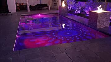 Une piscine dont le fond est illuminé par un motif géométrique, un espace détente abrité et un foyer.