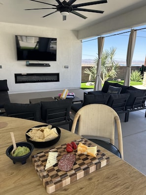 A modern living room with a flat-screen TV, a fireplace, a dining area with a wooden table, and a view of a garden and mountains.