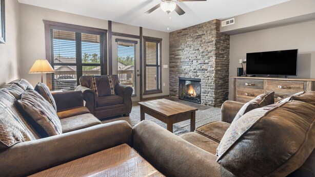 A living room with a stone fireplace, a ceiling fan, and a flat-screen TV. - 1 BDRM, Lake Community Condo near Silver Dollar City!  Ticket Deals and Resort Perks (Branson)