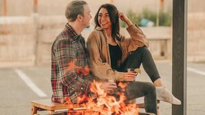 Una pareja sentada junto a una hoguera en un espacio al aire libre cubierto, con bancos y una estructura de madera rústica.