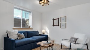 Una sala de estar con un sofá azul, un sillón blanco, una mesa de centro de madera y una mesita auxiliar.