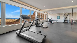 Ein großzügiger Fitnessraum mit Laufbändern, Ergometer und verschiedenen Trainingsgeräten. Große Fenster bieten Blick auf eine Küstenlandschaft.