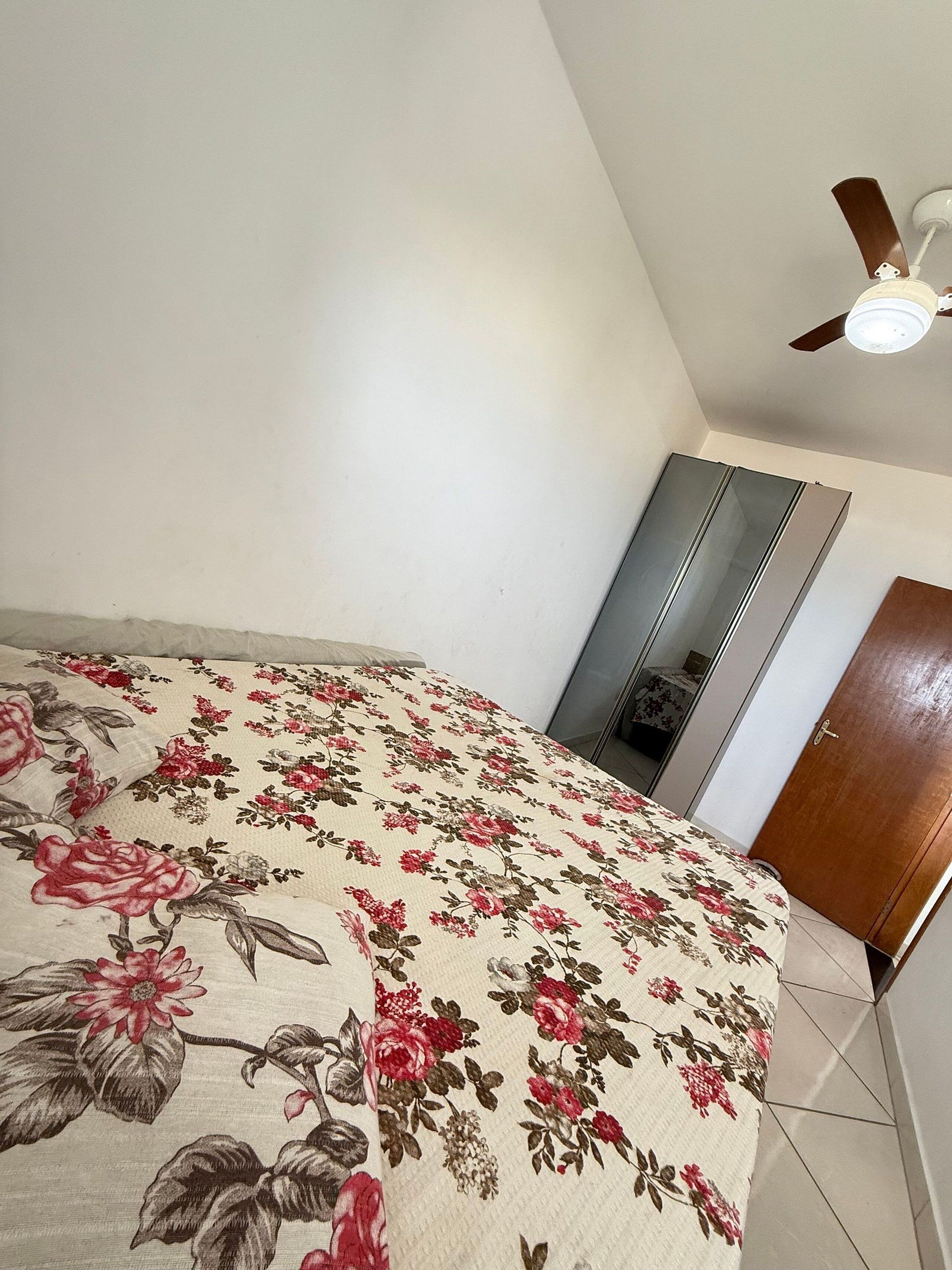 A bedroom with a floral bedspread, a ceiling fan, a wardrobe, and a door.