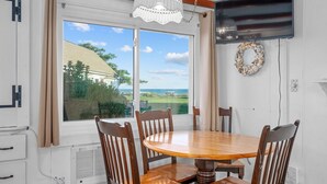 A dining area with a wooden table and chairs, a large window with a view of a house and greenery, and a decorative wreath on the wall. - Wake Up to Waves at Williams Port Cottage (South Yarmouth)