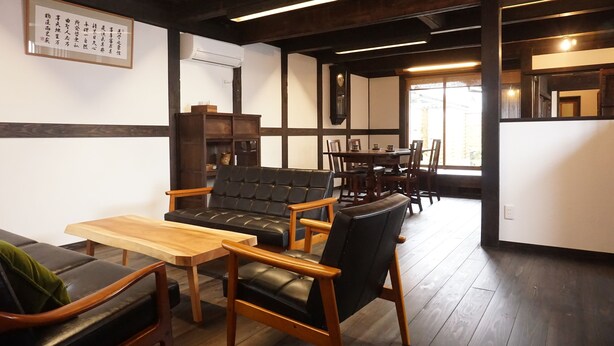 A modern living room with a black leather sofa, wooden chairs, and a wooden coffee table. There is a dining area with a table and chairs in the background. - Jisaburo Ozawa Residence (Takanabe)