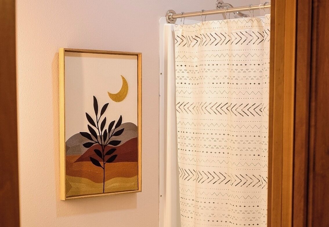 A bathroom with a framed artwork depicting a crescent moon and plant, and a patterned shower curtain.