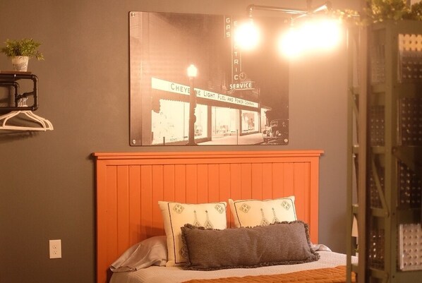Un dormitorio con cama, almohadas, una mesita de noche, un estante en la pared con una planta y un cartel de una estación de servicio.