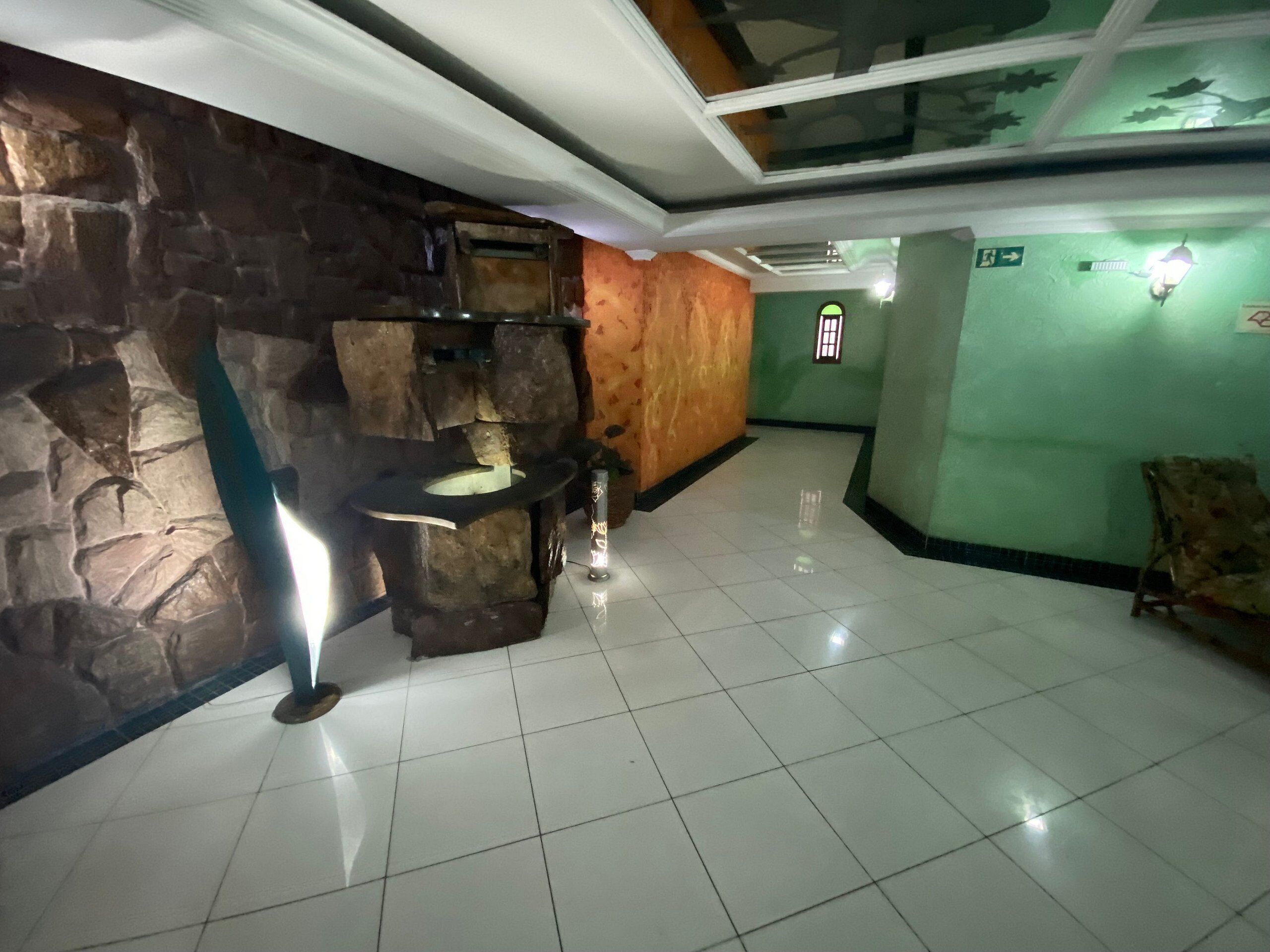 A lobby with a stone wall, a tiled floor, a small table with a lamp, and a green wall with a window.
