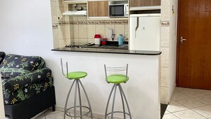 A compact kitchen with a microwave, stove, and refrigerator. There are two stools and a small table. - Pé Na Areia Praia da Ocean II (Praia Grande)