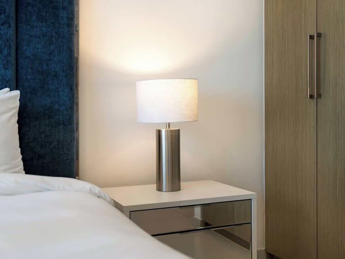 Ein ordentlich bezogenes Bett mit weißen Leinen, ein Nachttisch mit Lampe und ein Holzkleiderschrank.