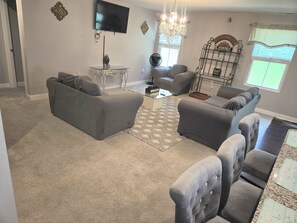 A living room with a grey sofa, a glass coffee table, a TV mounted on the wall, a chandelier, and a window with blinds. - Maison De La Paix: House of Peace (Charlotte)