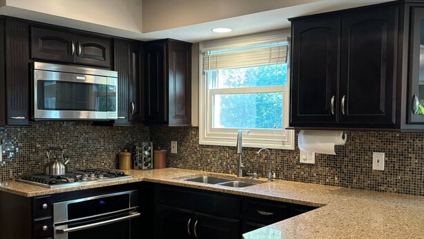 A modern kitchen with dark cabinetry, a stainless steel microwave, a built-in oven, granite countertops, and a window with blinds. - Luxury Modern Stay - 2000 Sqft (Indianapolis)